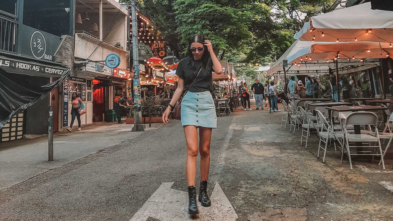 This image captures a lively street scene, featuring a young woman confidently walking down a pedestrian path lined with restaurants and bars. She is wearing a black top, a denim skirt, and boots, with string lights and outdoor seating adding to the vibrant atmosphere. The setting is shaded by trees and bustling with people, suggesting a trendy urban district.