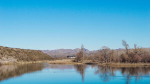 A serene landscape with a river reflecting a clear blue sky and leafless trees along the banks, suggesting it is either autumn or winter. In the background, low mountains and a structure with a dark roof, possibly a building or a rural cabin, can be seen. The image conveys a sense of tranquility in a natural setting.