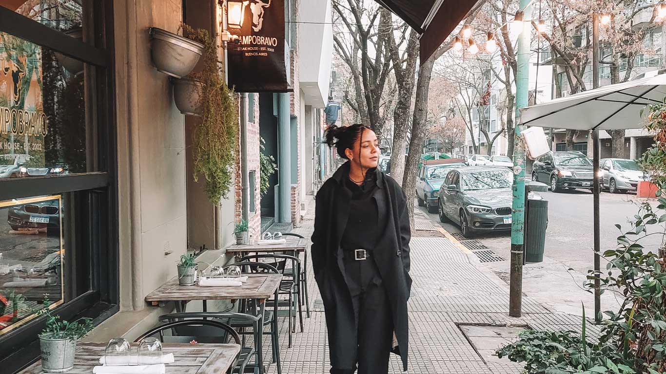 This image showcases a stylish street in Buenos Aires, Argentina, featuring a woman in dark attire walking past a cozy café with outdoor seating and plants. The scene captures the charm of the city’s tree-lined streets and boutique dining spots, ideal for exploring neighborhoods like Palermo. The mix of urban sophistication and casual ambiance highlights Buenos Aires’ vibrant culture.