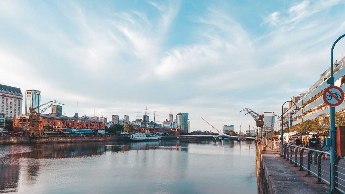 This image depicts Puerto Madero, a waterfront district in Buenos Aires, Argentina, featuring calm waters reflecting the surrounding buildings and a clear sky. The iconic Puente de la Mujer (Bridge of the Woman) is visible on the right, alongside modern architecture and industrial cranes adding character to the area. A no-cycling sign and promenade with people strolling emphasize the pedestrian-friendly ambiance.