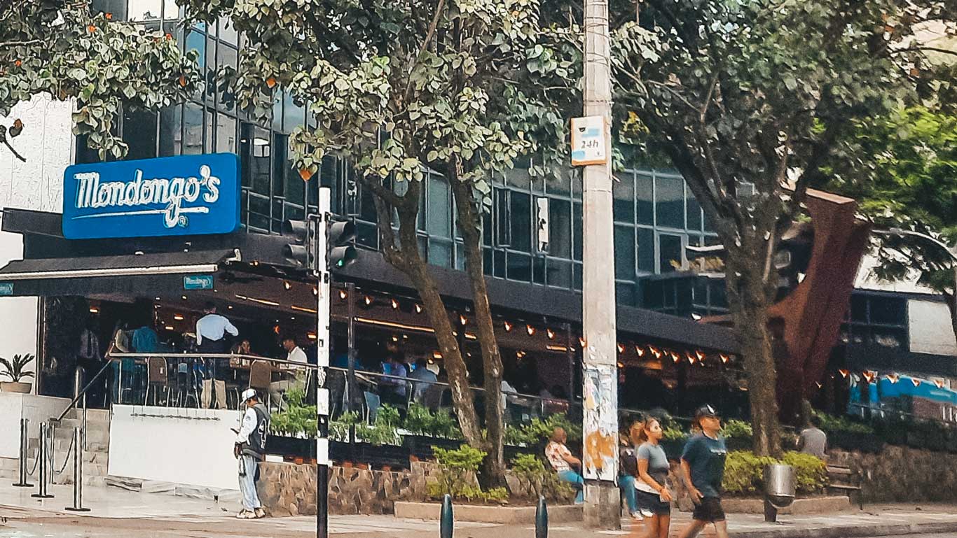 This image features the exterior of Mondongo's, a well-known restaurant with a bright blue sign and a sleek, modern design. The venue is bustling with activity, with people seated at tables inside and greenery enhancing the outdoor atmosphere. Trees and a busy street with passersby add a lively, urban vibe to the scene.