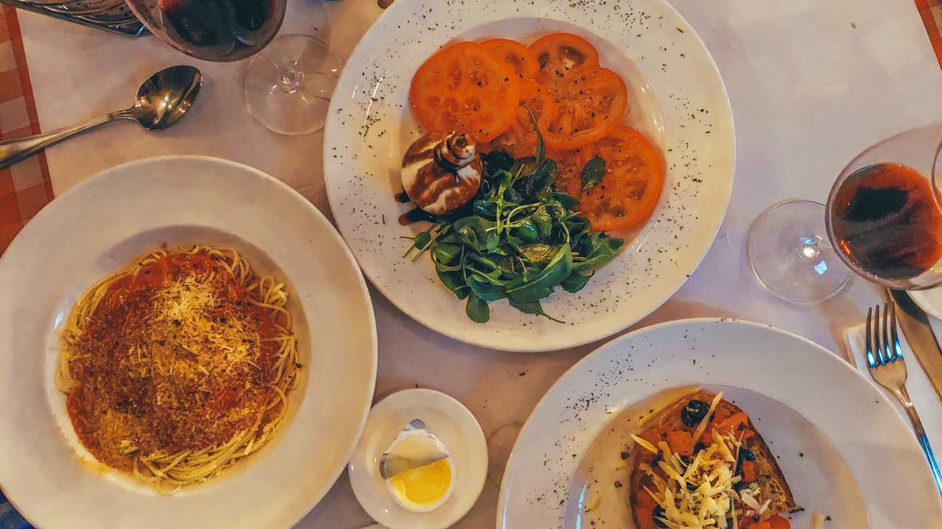 This image displays an elegant dinner table with three dishes arranged for a meal. One plate contains spaghetti topped with a rich tomato sauce and grated cheese, another features sliced tomatoes with a balsamic-glazed garnish and fresh greens, and the third shows a bruschetta-style dish with melted cheese and vegetables. Two glasses of red wine complement the meal, set on a white tablecloth for a cozy and upscale dining experience.