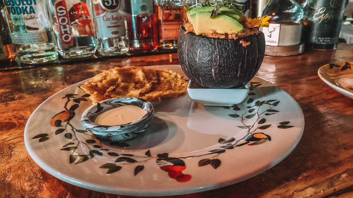 This image showcases a rustic dish presentation at a bar or restaurant. The plate includes a small black bowl filled with avocado slices and possibly a savory topping, accompanied by fried plantain slices and a dipping sauce, all set against a decorative ceramic plate. Behind the dish, there’s a lineup of liquor bottles, hinting at a vibrant bar setting.