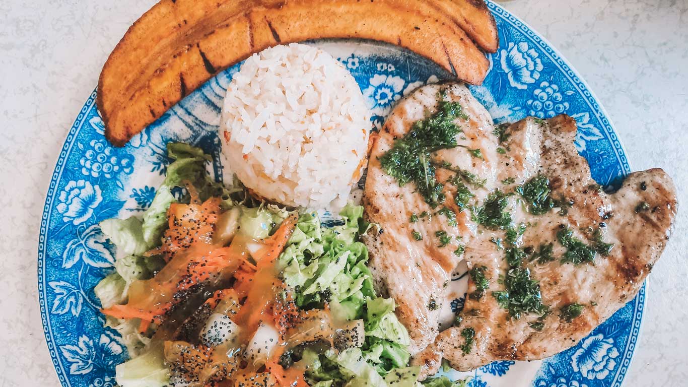 This image shows a traditional meal served on a decorative blue plate. The dish includes grilled meat garnished with fresh herbs, a portion of white rice, crispy fried plantains, and a fresh salad with lettuce, carrots, and a light dressing. The vibrant colors and balanced presentation highlight a comforting and wholesome meal.