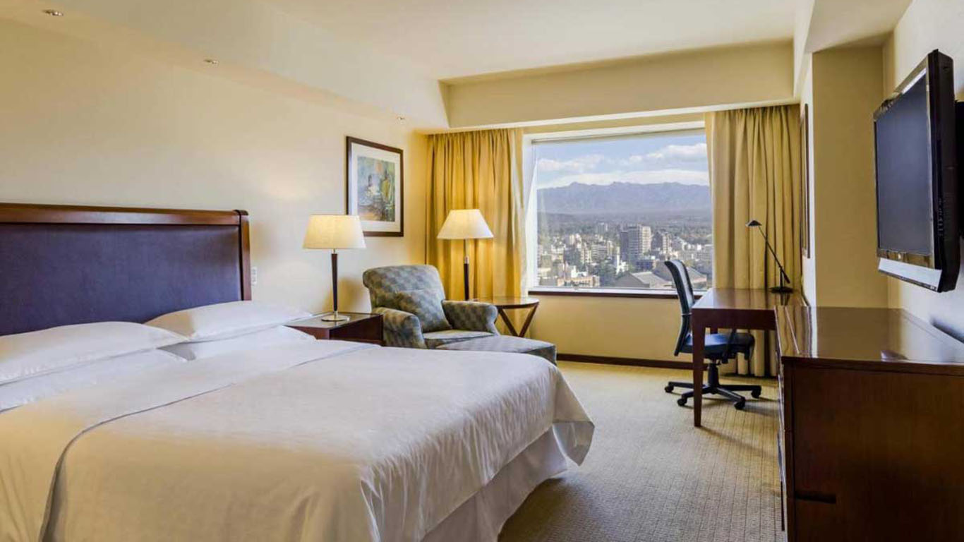 This image shows a hotel room with a spacious and well-lit design. It features a large bed with white linens, a seating area with an armchair, a wooden desk with a chair, and a mounted flat-screen TV. The window provides a view of a cityscape and distant mountains, framed by beige curtains. 