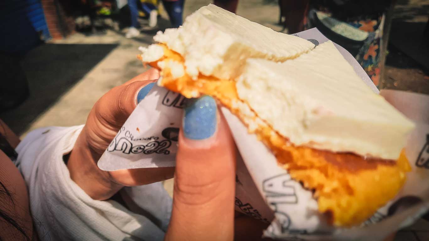 This image features a close-up of a hand holding a piece of traditional Colombian arepa, topped with a generous slice of fresh white cheese. The arepa has a golden, crispy texture, contrasting with the creamy, soft cheese. The setting appears casual and outdoor, highlighting street food or a local snack experience.