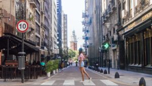 This image shows a lively street in Buenos Aires, Argentina, featuring tall buildings, a 10 km/h traffic sign, and a pedestrian walking across a crosswalk. In the distance, the Torre Monumental (Clock Tower) is visible, adding a historical charm to the scene. Street cafés and greenery enhance the urban vibe.