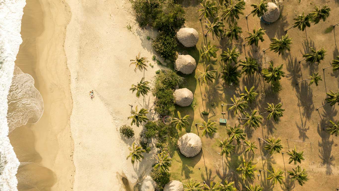 This image depicts an aerial view of a tropical coastline, likely Tayrona National Park. On the left, golden sandy beaches meet gentle ocean waves, while on the right, clusters of palm trees and thatched-roof huts create a serene landscape. This balance of lush greenery and pristine shoreline highlights the area's natural beauty.