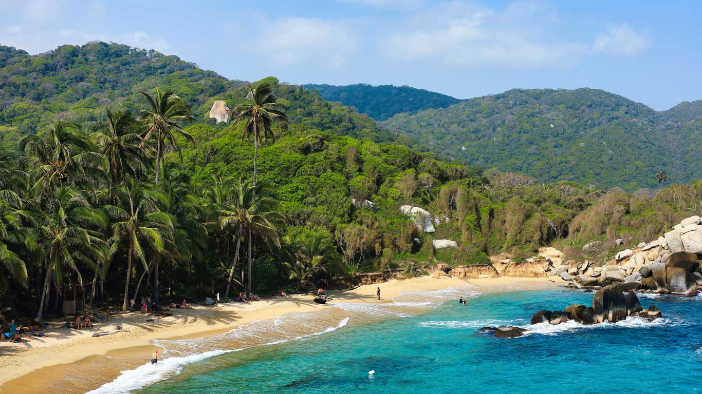 This image shows a picturesque beach in Tayrona National Park, with crystal clear turquoise waters meeting a golden sandy shore lined with lush palm trees. In the background, lush green hills create a dramatic backdrop, emphasizing the tropical and serene atmosphere. This area with beaches is an excellent choice for where to stay in Tayrona National Park.