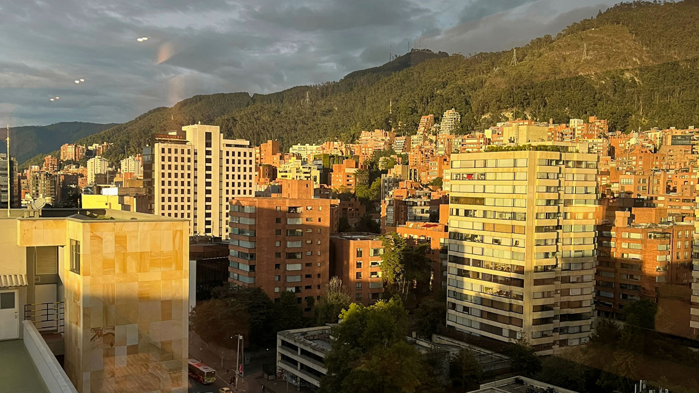 A vibrant view of Chapinero, Bogotá, with modern apartment buildings bathed in golden sunlight against the lush, green backdrop of the Andes mountains. The red-brick and cream-colored high-rises reflect the city's blend of contemporary living and natural beauty. 