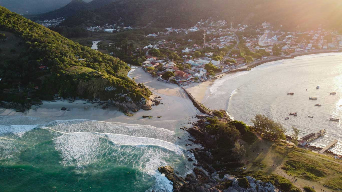 Aerial view of Armação Beach in Florianópolis at sunset, featuring a curved shoreline, lush green hills, and a charming coastal village with white and red-roofed houses. Waves crash onto the sandy beach, while small boats float in the bay, offering a picturesque destination