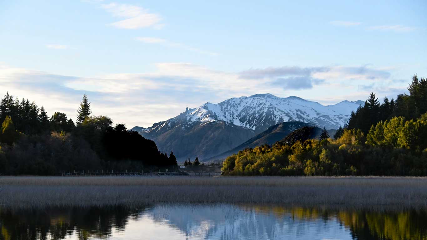 A tranquil lake reflecting the surrounding greenery and snow-capped mountains in Bariloche, Argentina. The scene is framed by dense forests on either side, with the peaks of the Andes mountains rising dramatically in the background. The sky is softly lit, creating a serene and picturesque landscape.