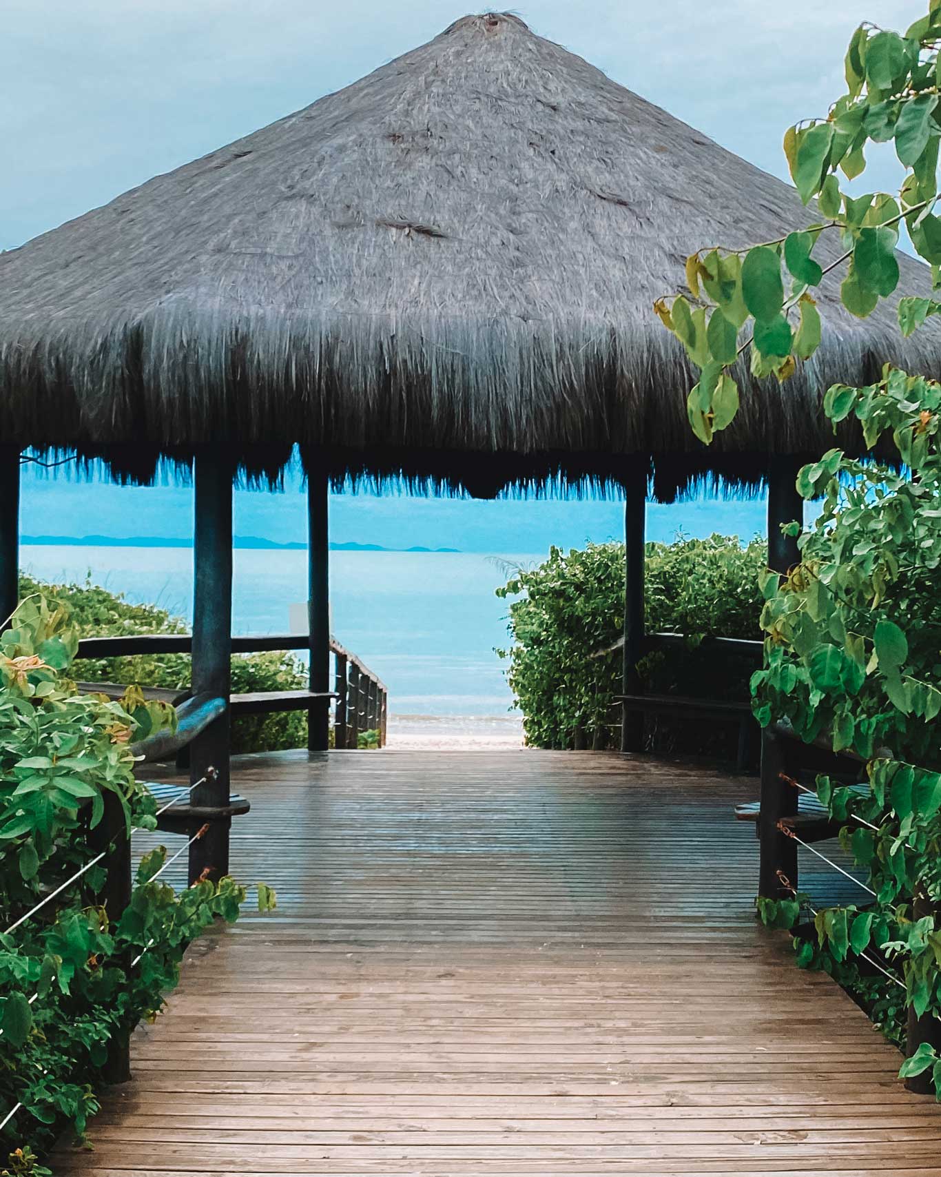 Wooden boardwalk leading to a beachfront cabana with a thatched roof, surrounded by lush green foliage and overlooking the serene blue ocean. This tranquil spot captures the tropical charm of the best place to stay in Florianópolis, offering relaxation and stunning sea views.