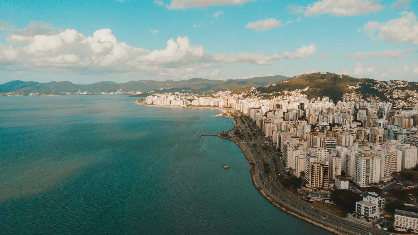 A coastal view of Florianópolis, Brazil, showcasing a skyline of modern high-rise buildings along the shoreline with mountains in the background. The clear blue water of the bay meets a winding road that hugs the coast, under a bright sky with fluffy clouds.