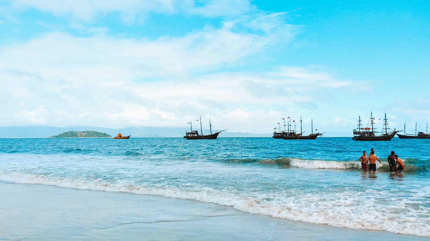 Beach scene at Canasvieiras in Florianópolis, featuring turquoise waters, people enjoying the surf, and several pirate-themed boats anchored offshore.