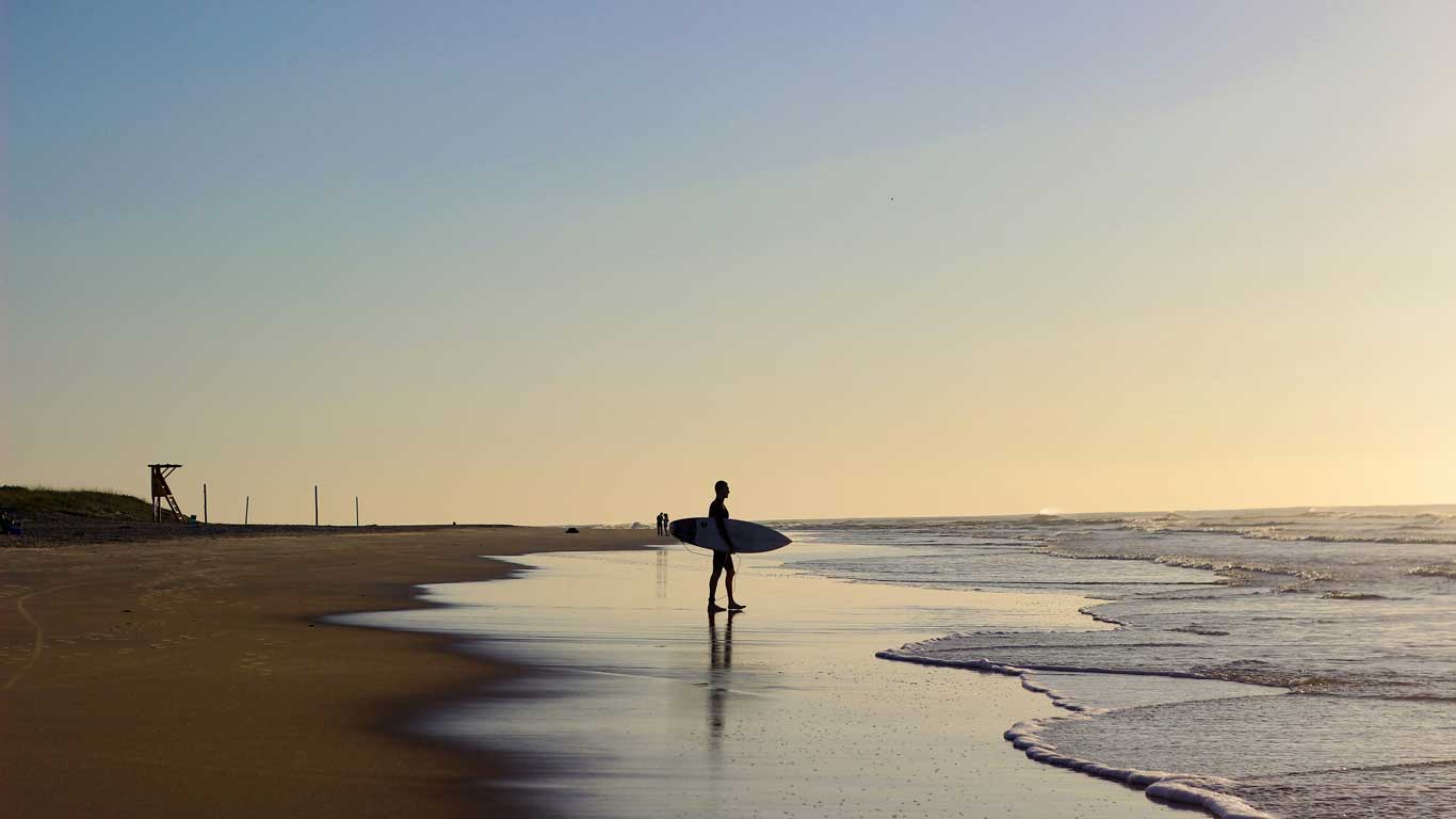 Sunset scene at a tranquil beach in Florianópolis, featuring a lone surfer with a board standing at the water's edge.