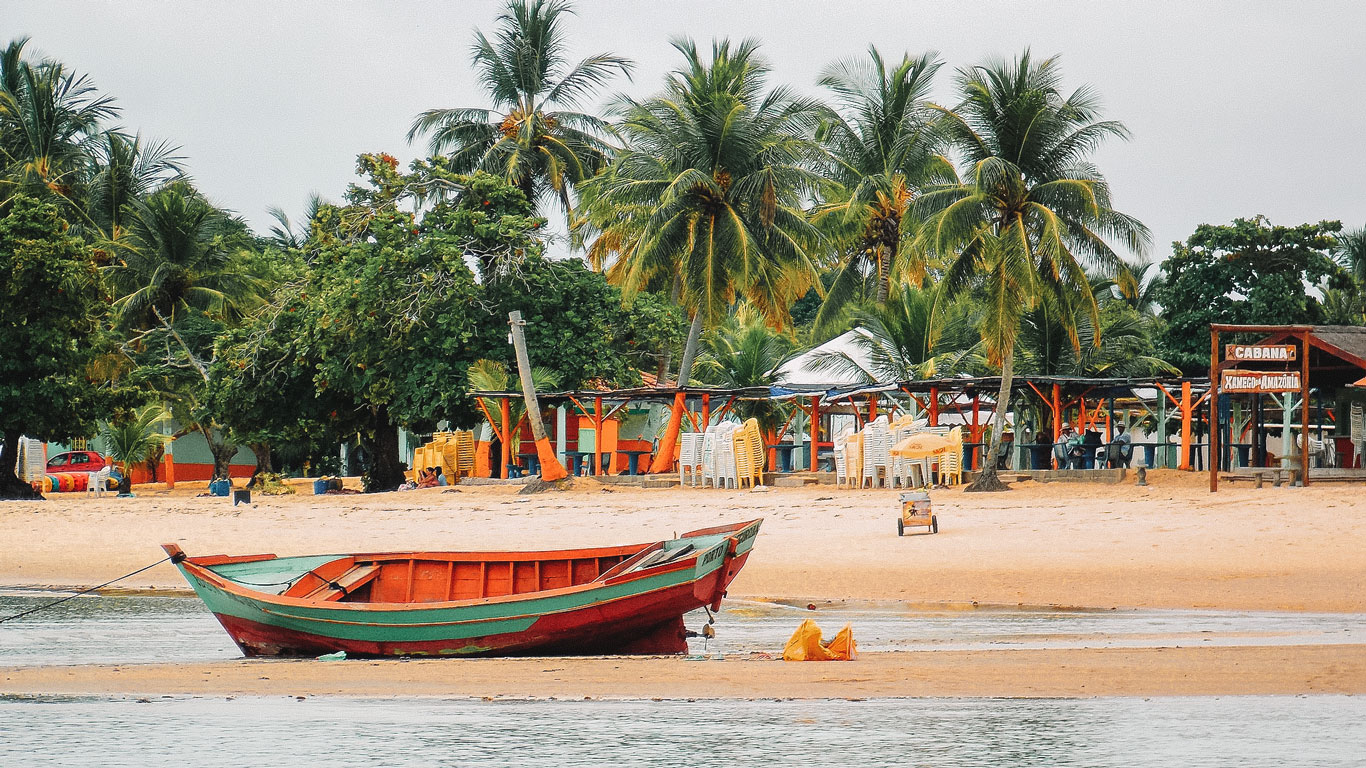 Coroa Vermelha Beach featuring a colorful wooden boat resting on the sand, surrounded by tall palm trees and beachside cabanas. The vibrant scene includes outdoor seating areas, a relaxed atmosphere, and lush greenery, making it a picturesque destination in Porto Seguro