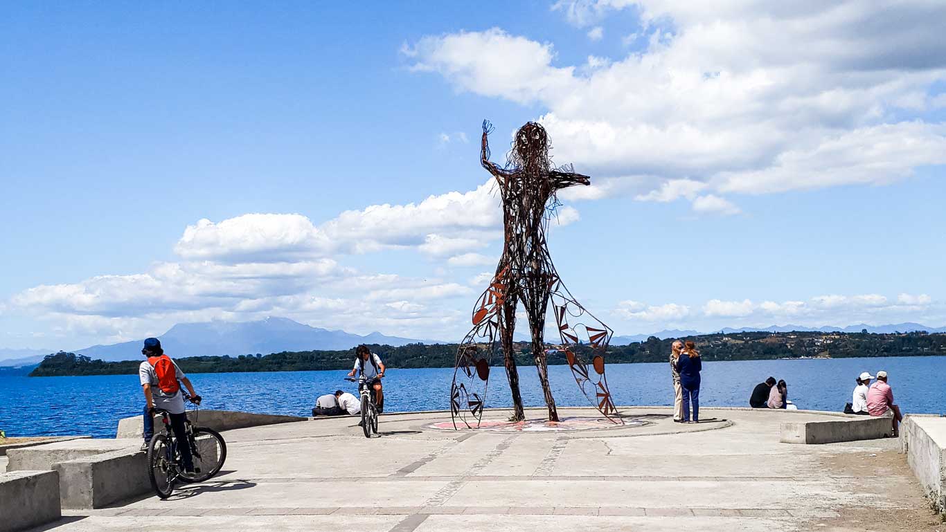 A waterfront scene in Puerto Varas, Chile, featuring a tall metal sculpture of a figure with one arm raised, standing on a concrete pier. Cyclists, pedestrians, and groups of people are enjoying the sunny day, with the lake and distant mountains visible under a partly cloudy sky. The blue waters reflect the serene atmosphere of the lakeside setting.