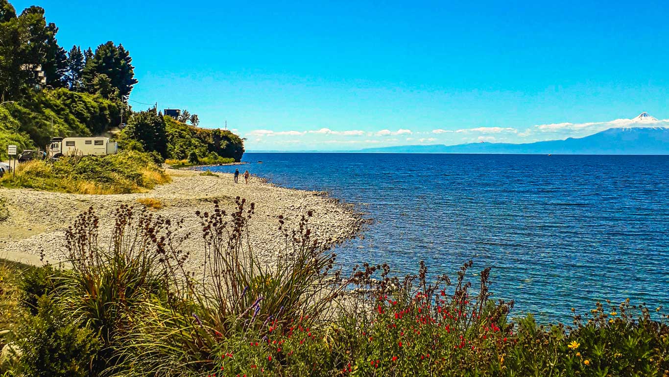 A pebble beach along the coast of Puerto Varas, Chile, with vibrant green foliage and red flowers in the foreground. The blue waters of the lake stretch out to a clear horizon, with a snow-capped volcano visible in the background. A couple of people walk along the shore near parked vehicles and lush greenery under a bright blue sky.