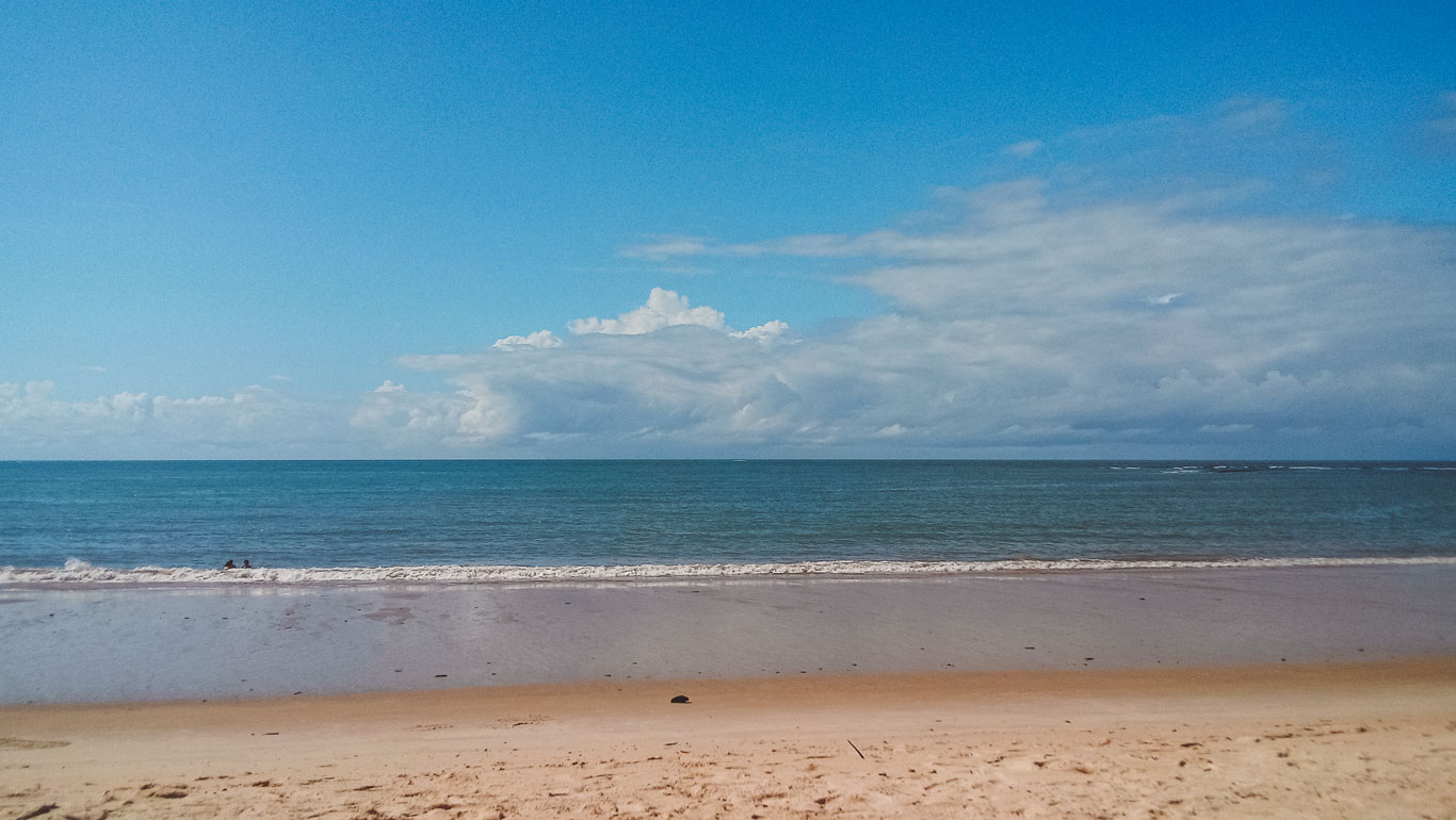 uruípe Beach in Porto Seguro, featuring a pristine stretch of sand meeting calm, blue waters under a partly cloudy sky. A peaceful shoreline perfect for relaxation and enjoying the natural beauty of this coastal destination."