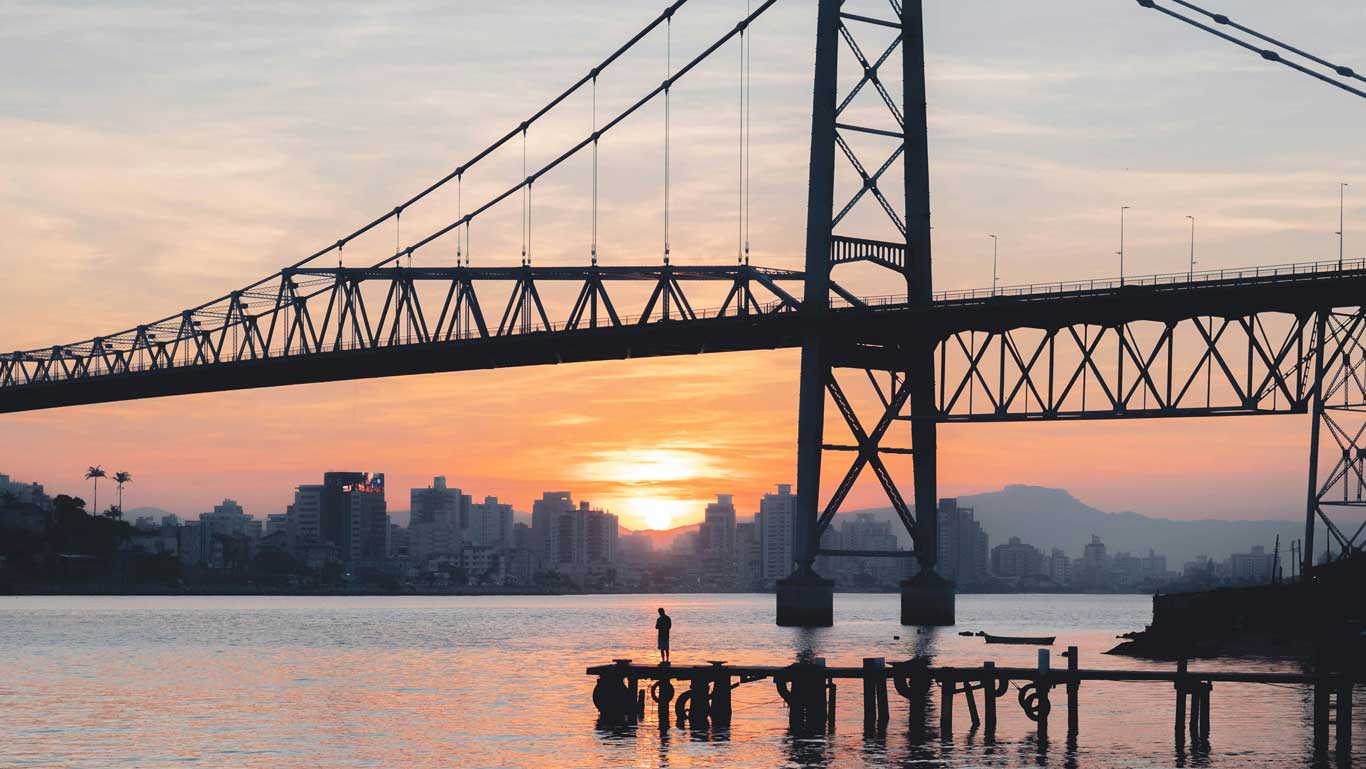 Silhouette of the Hercílio Luz Bridge during sunset in Florianópolis, with a person standing on a rustic wooden pier and the city's skyline in the background. This iconic view highlights the charm of downtown, one of the many vibrant places to stay in Florianópolis.