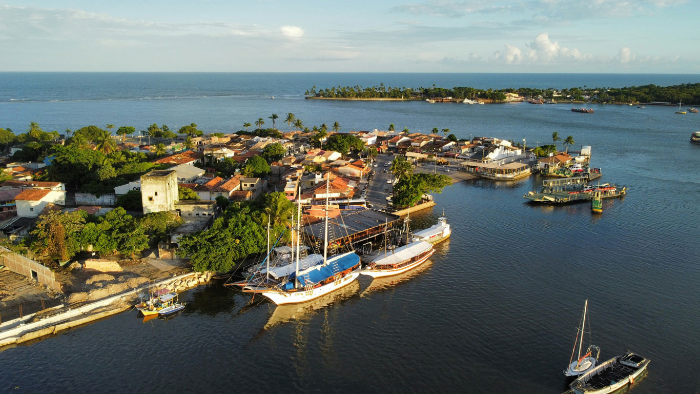 This image shows a scenic aerial view of a coastal town, featuring vibrant buildings with red-tiled roofs surrounded by lush greenery. Several boats and yachts are docked at a calm waterfront, with a small island visible in the background under a clear blue sky. The image captures the charm and tranquility of a seaside Porto Seguro community.