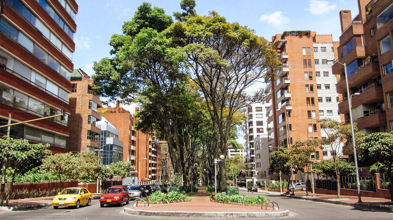 A tree-lined street in Bogotá's upscale El Chicó neighborhood, showcasing lush greenery and modern apartment buildings. The bright blue sky, red-brick façades, and pedestrian-friendly pathways highlight this area as one of the best places to stay in Bogotá for a mix of nature and city living.