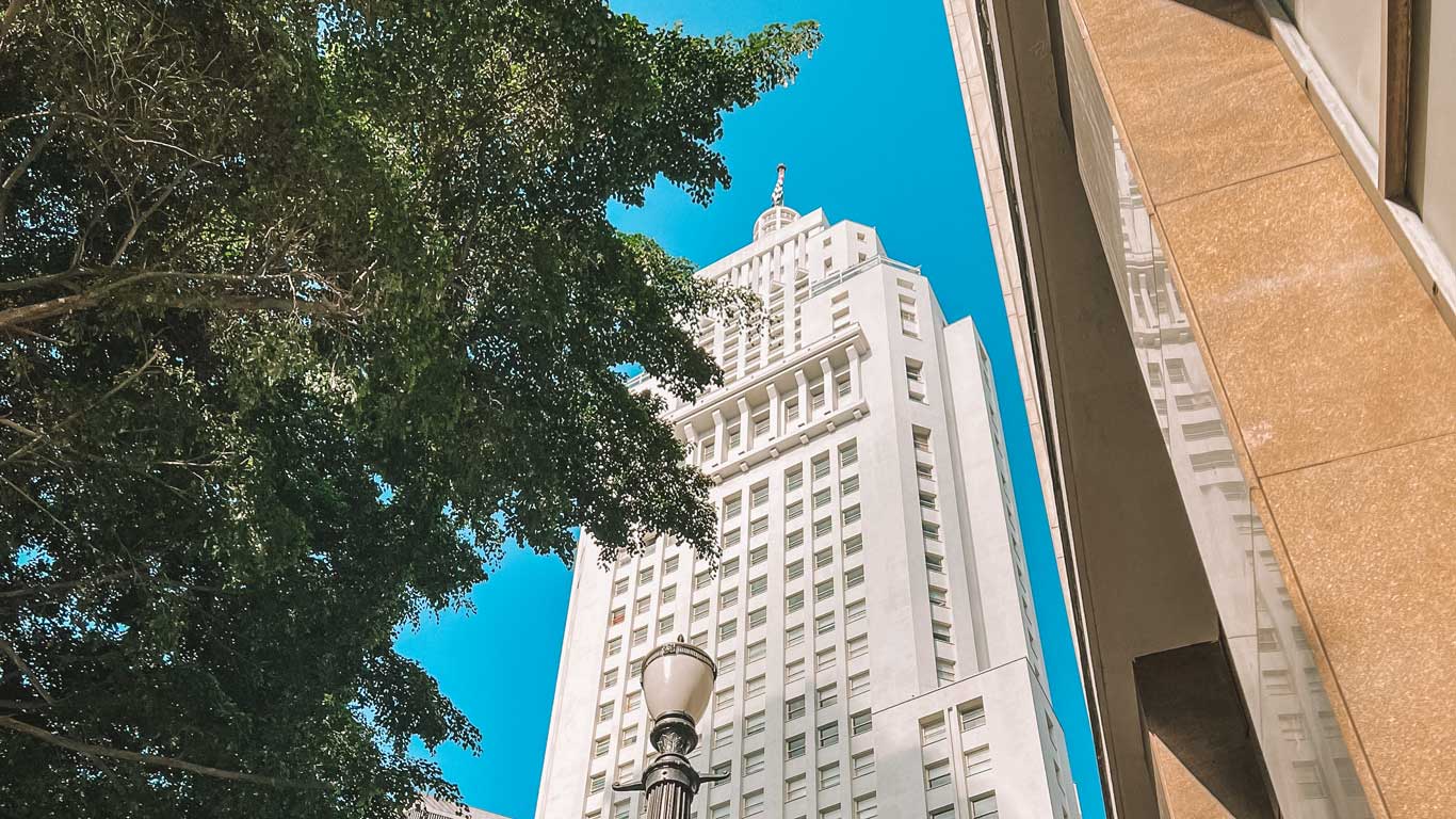 An upward view of the iconic Farol Santander building in São Paulo, framed by tree branches and modern architecture, under a bright blue sky.