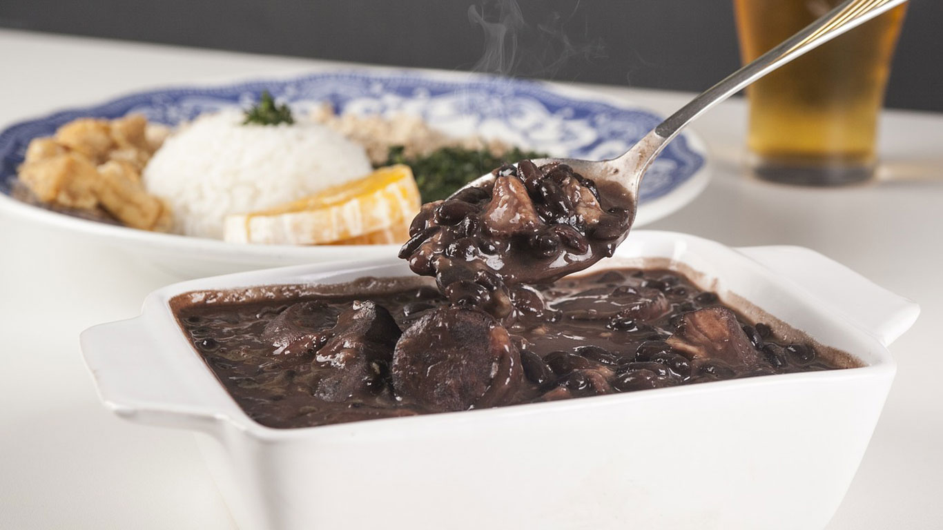 A steaming bowl of feijoada, a traditional Brazilian black bean stew with chunks of meat, served alongside rice, collard greens, orange slices, and fried bananas on a plate in the background.