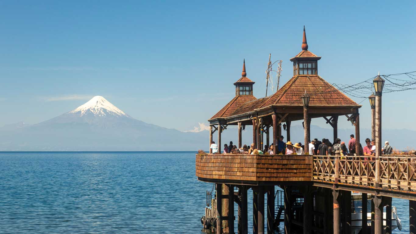 A wooden pier in Frutillar, Chile, with a group of people enjoying the lakeside view under a gazebo-style structure. The scene overlooks the calm blue waters of the lake, with a snow-capped Osorno Volcano in the background beneath a clear blue sky. String lights and lampposts line the pier, adding charm to this picturesque setting.