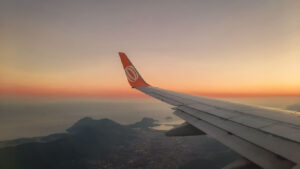 Image of an airplane wing with the airline's logo in orange at the tip, seen during a flight at sunrise or sunset. Below, a coastal landscape with mountains and islands can be seen, under a sky with shades of orange and yellow near the horizon.