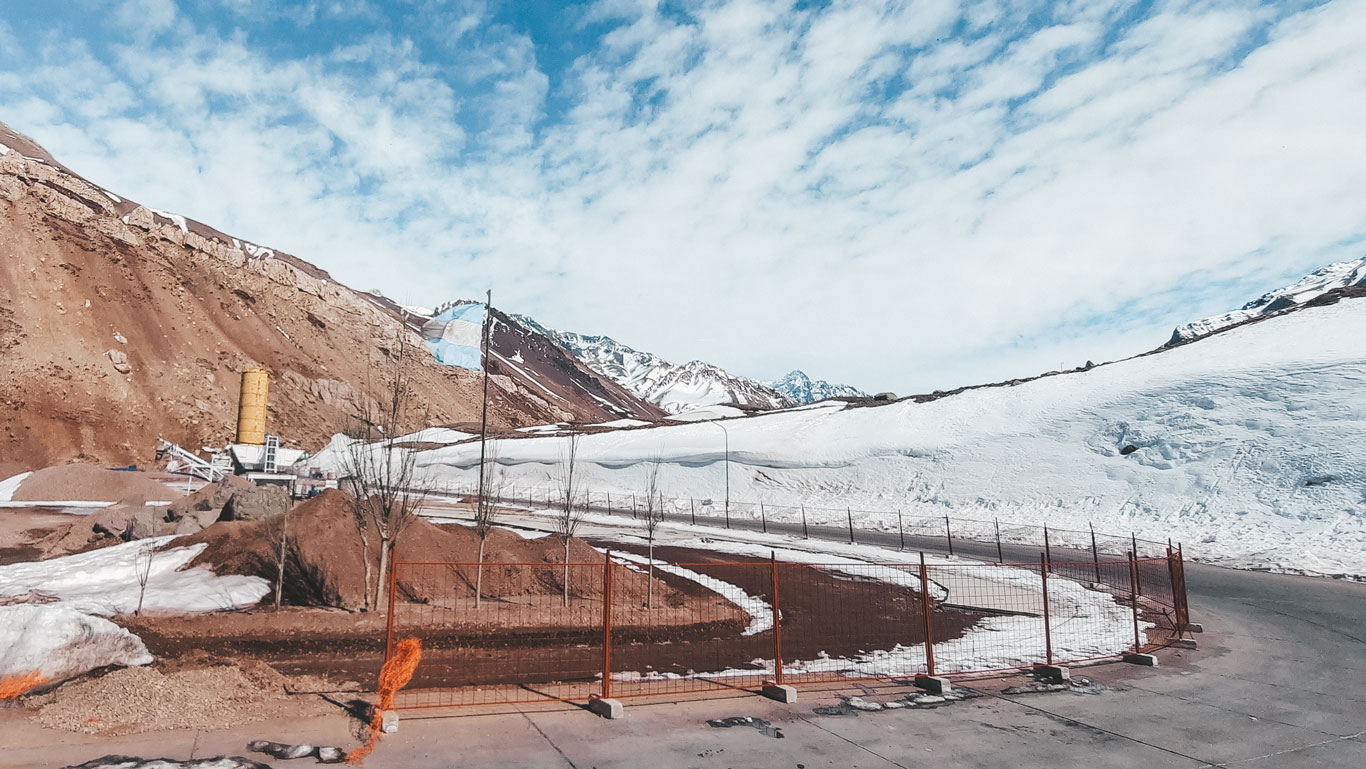 The image shows a mountain road partially surrounded by snow, with rocky hills and snow-capped mountains in the background. The blue sky with scattered clouds complements the cold and arid landscape, and an industrial structure with a large yellow tank is visible to the left, near the road.