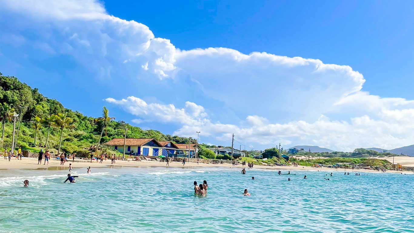 View of Ingleses Beach in Florianópolis, with turquoise waters and people enjoying the ocean and sandy shore. The backdrop features lush greenery, colorful beachfront houses, and a vibrant atmosphere, showcasing an ideal spot for those seeking where to stay in Florianópolis.