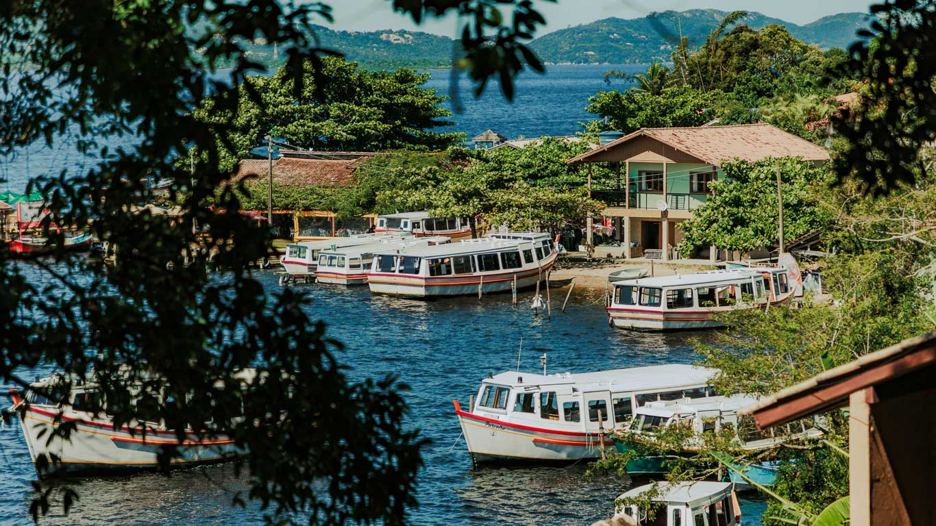 View of Lagoa da Conceição in Florianópolis, featuring a tranquil harbor with several white boats docked near a green two-story building surrounded by lush foliage. The blue waters and distant mountains highlight the charm of this popular spot, ideal for those searching where to stay in Florianópolis.