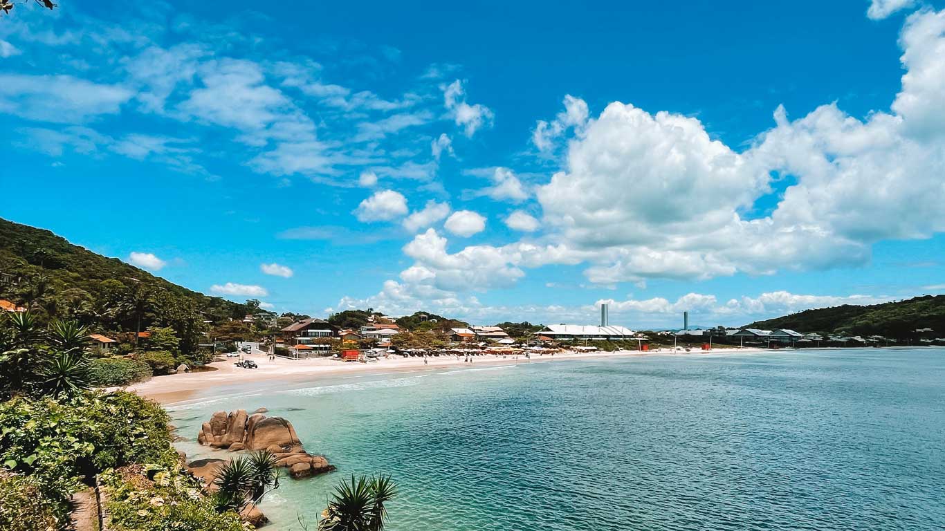 Scenic view of Lagoinha Beach in Florianópolis, featuring clear turquoise waters, soft sandy shores, and a backdrop of green hills and colorful beach houses. 