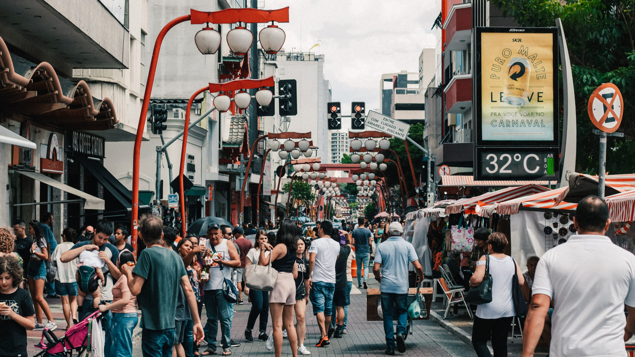 A bustling street scene in São Paulo's Liberdade neighborhood, featuring red Japanese-style lanterns, market stalls, and crowds of people enjoying the vibrant atmosphere.