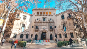 This image shows an ornate building with intricate carvings and architectural details under a clear blue sky in Mendoza. The structure has signs reading "Banco Hipotecario Nacional" and features two Argentine flags on the facade. In front of the building, a colorful sign spelling out "CULTURA" is displayed. Several people are walking along the tree-lined sidewalk.