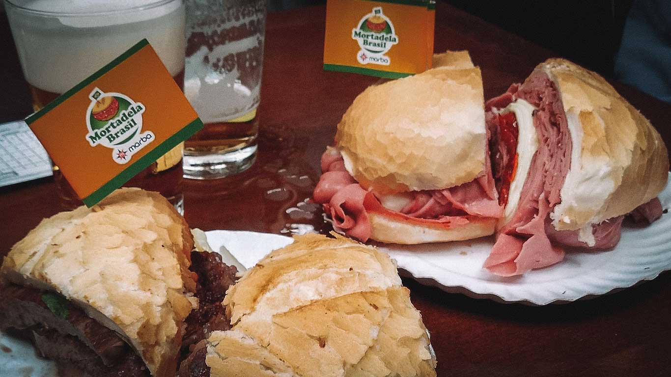 A close-up of two iconic mortadella sandwiches served on crusty bread, accompanied by drinks, with "Mortadela Brasil" flags marking the meal. This classic culinary experience is a must-try at the Mercado Municipal.