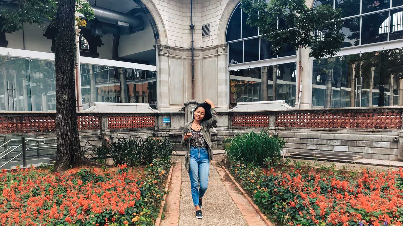 A woman stands confidently in a garden filled with vibrant red flowers in front of the ornate facade of the Catavento Museum in São Paulo. 