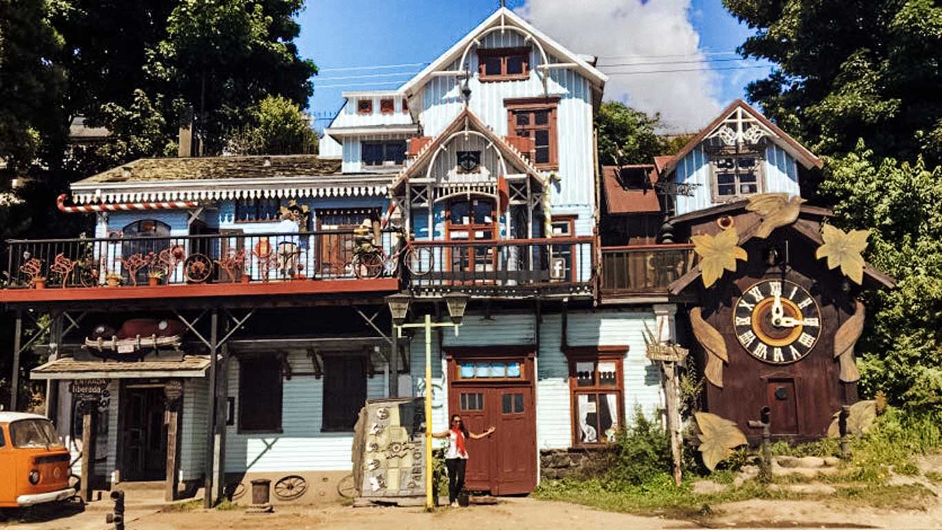 The Pablo Fierro Museum in Puerto Varas, Chile, a whimsical, multi-story wooden building with eclectic decorations like bicycles, old signs, and a large clock adorned with maple leaves. A person stands in front of the entrance beneath the clock, with an orange vintage van parked nearby. This artistic landmark is surrounded by trees and offers a unique cultural stop for visitors exploring things to do in Puerto Varas.