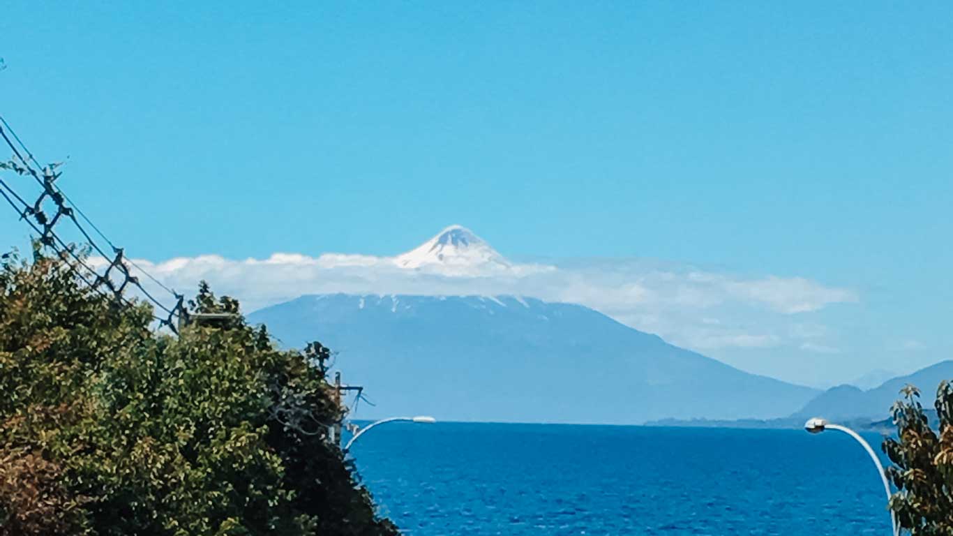 A view of the Osorno Volcano in Chile, with its snow-capped peak rising above a blanket of clouds. The foreground features blue lake waters, lush green trees, power lines, and street lamps. The volcano's symmetrical cone stands tall against a clear blue sky, creating a striking landscape.