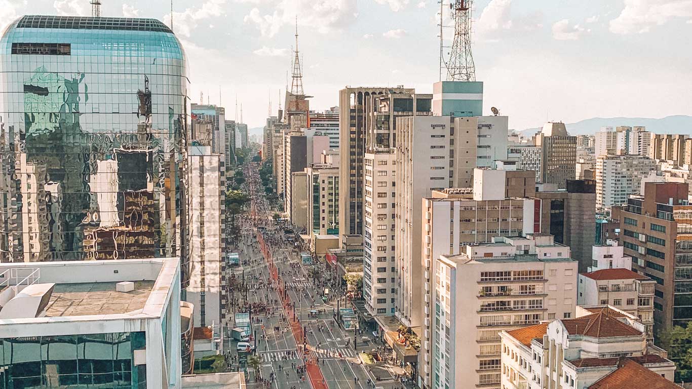 This image shows a high-angle view of Paulista Avenue in São Paulo, Brazil, with tall modern buildings on either side and reflective glass facades capturing the urban skyline. The avenue is bustling with people and vehicles, and the red bus lane running down the center adds a pop of color to the scene.