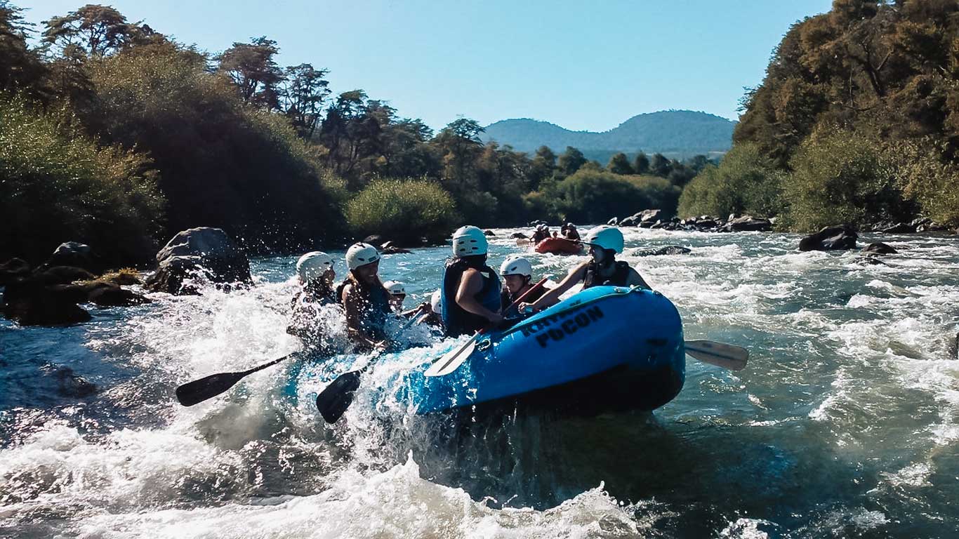 A group of people in helmets and life jackets navigate white-water rapids in a blue inflatable raft marked "Pucón." The river is surrounded by lush green trees and rolling hills, with water splashing around the raft as they paddle. This exhilarating scene captures the thrill of rafting in Pucón, Chile, known for its adventure sports and natural beauty.