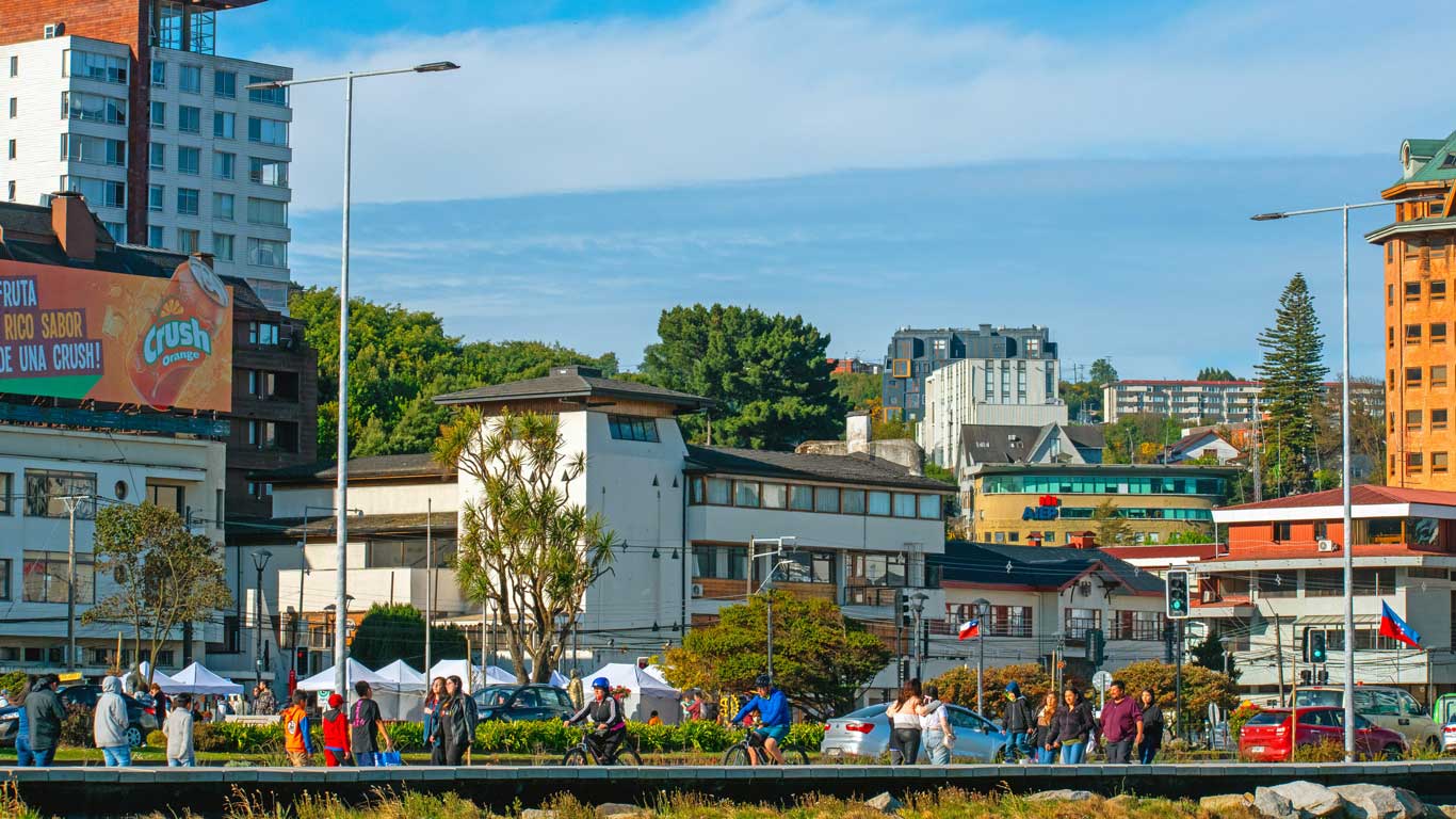 A bustling street scene in Puerto Montt, Chile, featuring pedestrians, cyclists, and cars along the waterfront. Various modern buildings, including a tall apartment complex and commercial establishments, are visible in the background.