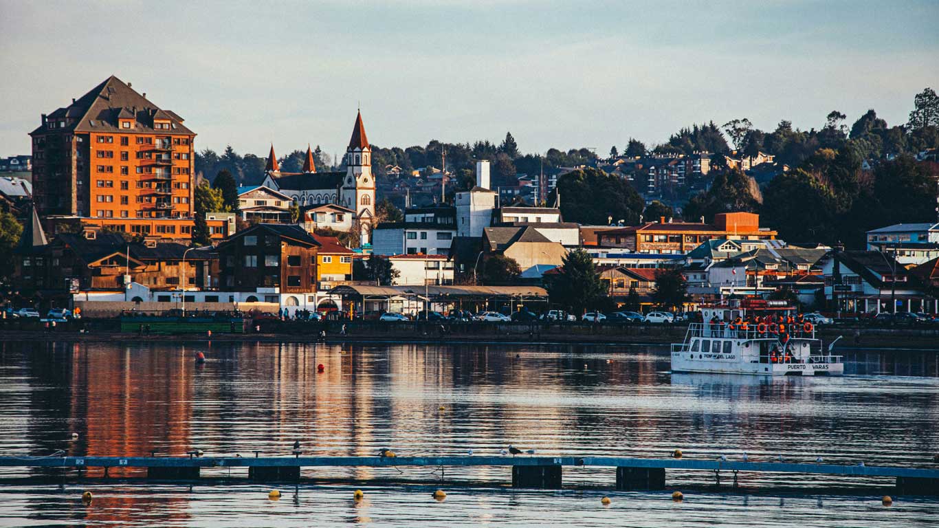 A scenic view of Puerto Varas, Chile, featuring a white and red boat on the calm lake in the foreground. The background showcases a variety of buildings, including a prominent church with red spires, surrounded by modern and traditional architecture.