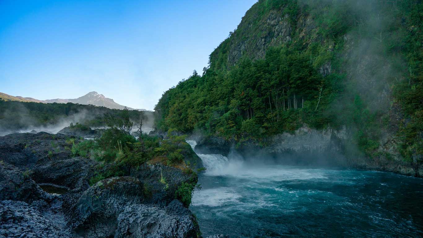  The Saltos de Petrohué waterfalls in Chile, with powerful turquoise waters rushing through volcanic rock formations. Mist rises from the falls, surrounded by dense green forests and a steep cliff on the right side. A snow-capped mountain peak is visible in the distance under a clear blue sky, making this a stunning natural attraction near Puerto Varas.