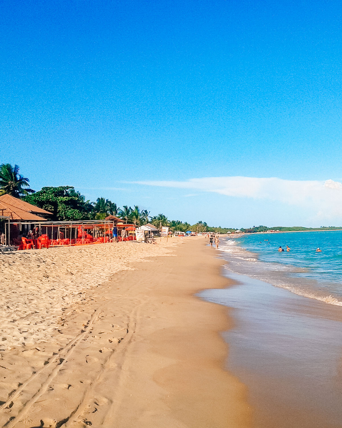 This image shows sun-drenched Taperapuã Beach with golden sand stretching along the coastline, surrounded by calm, turquoise waters. Lined with beachfront cabanas and bright red chairs under umbrellas, it features people enjoying the waves and strolling along the shore. The backdrop includes lush greenery and a vibrant blue sky, showcasing a lively tropical paradise.