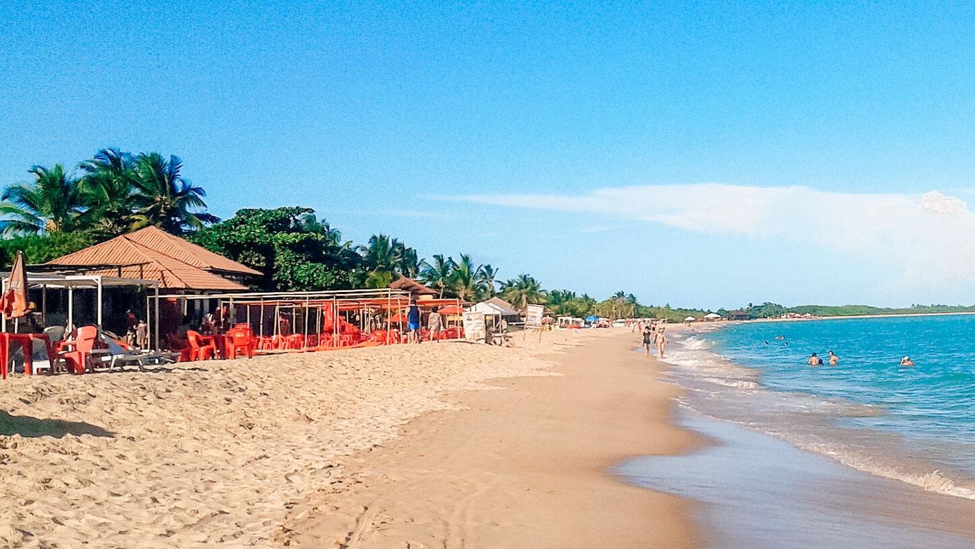 This image shows sun-drenched Taperapuã Beach with golden sand stretching along the coastline, surrounded by calm, turquoise waters. Lined with beachfront cabanas and bright red chairs under umbrellas, it features people enjoying the waves and strolling along the shore. The backdrop includes lush greenery and a vibrant blue sky, showcasing a lively tropical paradise.