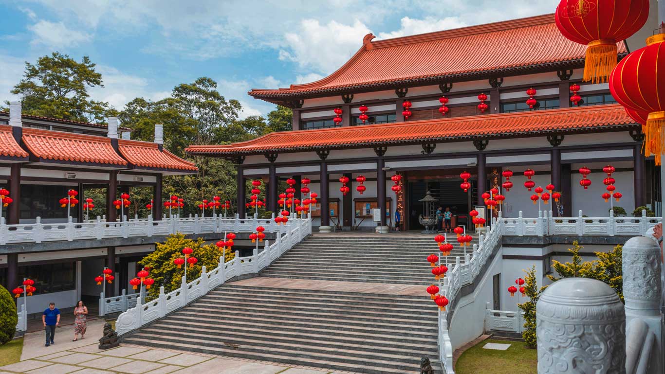 The Zu Lai Temple in São Paulo, featuring traditional Chinese architecture with red-tiled roofs, red lanterns, and intricate white railings, surrounded by lush greenery. Visiting this tranquil Buddhist temple is one of the most peaceful and cultural things to do in São Paulo.