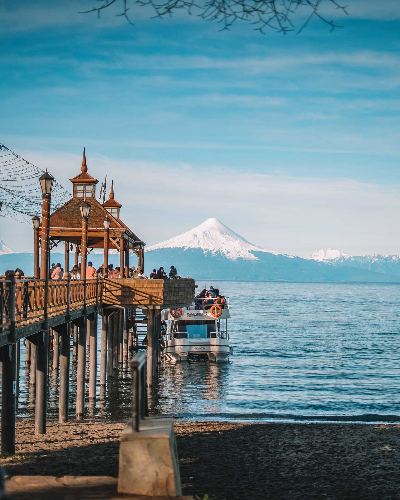 A wooden pier in Puerto Varas, Chile, with a crowd of people standing beneath decorative lanterns and string lights. A boat approaches the pier on the calm blue water, while a snow-capped volcano dominates the background beneath a clear sky. The sandy shoreline and reflections of the pier are visible in the foreground.